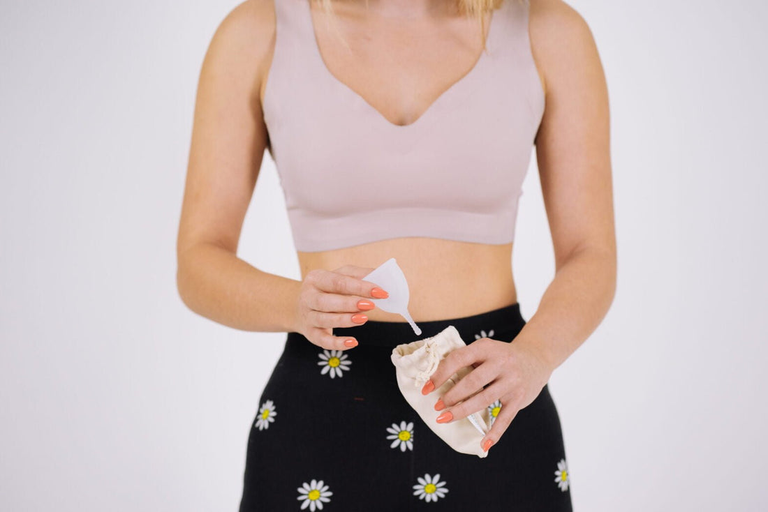 Femeie ținând o cupă menstruală albă și un săculeț textil reutilizabil, simbol al menstruației sustenabile.